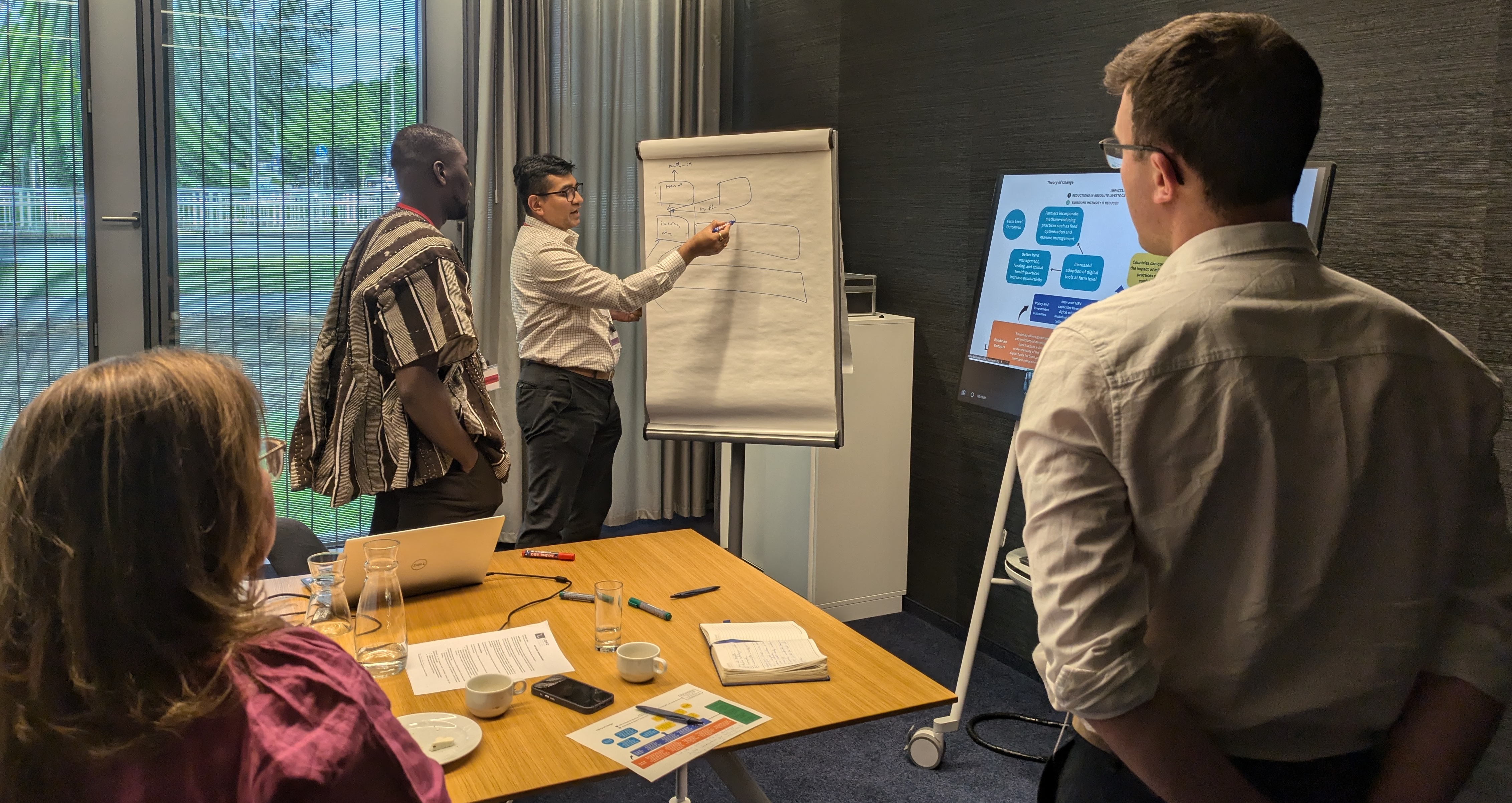 Four people stand in a meeting room, looking at a person who is presenting a diagram on a flipchart. Between them is a table strewn with notebooks, papers, pens, laptops and coffee cups.