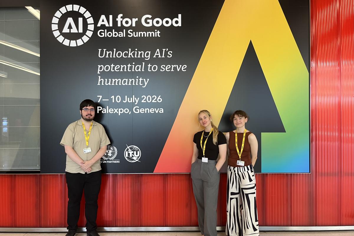 Three adults stand in front of a large banner that reads AI for Good with a large multicolour stylised letter A against a black background.