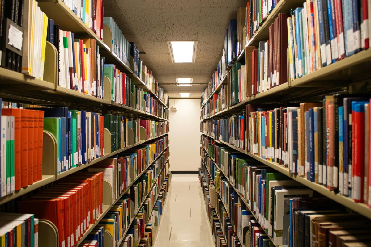 A view between two long rows of books in a library with a door at the end of the row. There is an overhead fluorescent light on the ceiling. 