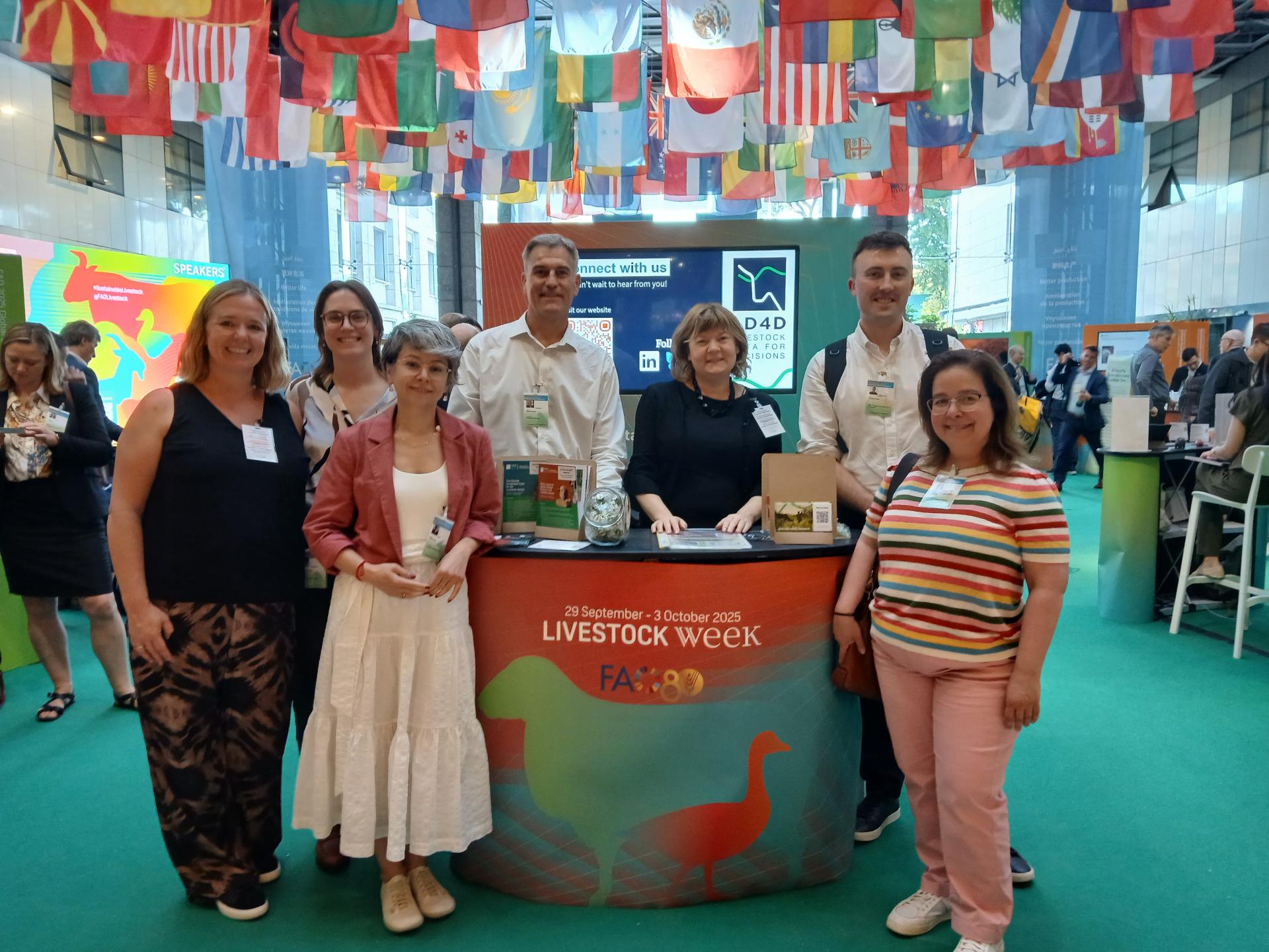 A group of adults stand around an exhibition stand, which is decorated with an orange, green and blue illustration of livestock in silhouette. The sign reads "FAO Livestock Week 2025". From the ceiling hang numerous flags from countries around the world.