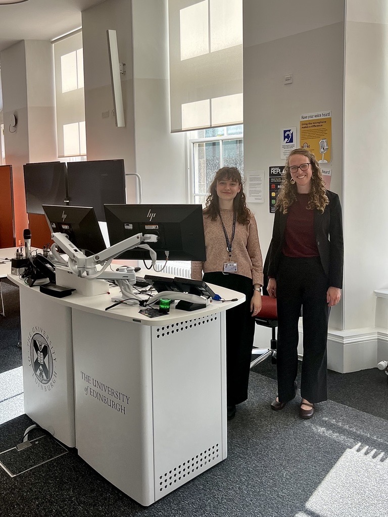 Two women stand smiling behind a podium with computer monitors on it.